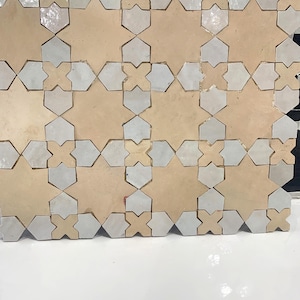 Puede incluir: Primer plano de una pared enlosada con un patrón geométrico de azulejos de cerámica beige y blanco. Los azulejos están dispuestos en un patrón repetitivo de estrellas y cruces.
