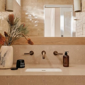 May include: A modern bathroom vanity with a light brown countertop, a white rectangular sink, and bronze fixtures. A brown soap dispenser and a black candle sit on the counter. A vase with dried flowers and a mirror are in the background.