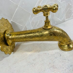 May include: A gold-toned faucet with an ornate design. The faucet has a cross-shaped handle and a spout that curves out and down. The faucet is mounted on a wall.