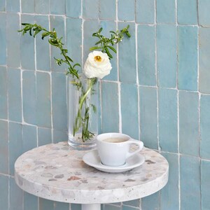 May include: A small, round table with a speckled top holds a white teacup and saucer, and a clear vase with a white flower and greenery. The table has a white base and is set against a light blue tiled wall.