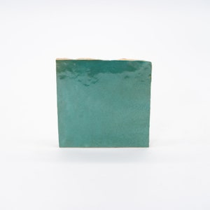 May include: A square tile with a teal green glaze. The tile has a slightly rough texture and is slightly chipped on the edges.