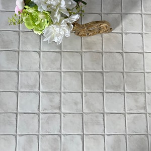 May include: A gold-colored decorative bracelet with an open design rests on a textured, square-tiled surface. A bouquet of white, green, and pink artificial flowers is in the upper left corner. The background is a neutral, off-white color.
