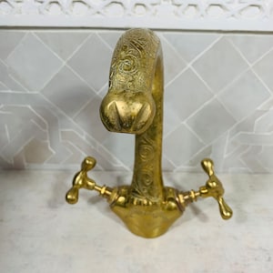 May include: A vintage brass faucet with an ornate design. The faucet features a curved spout and two cross-shaped handles. The brass has a warm, aged patina, and the spout is decorated with etched floral patterns.