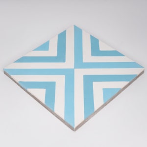 May include: Square tile with a geometric design. The tile features a pattern of alternating light blue and white lines, creating a cross-like design. The tile is square-shaped and has a light brown edge.