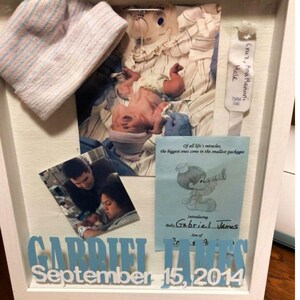 Könnte beinhalten: Ein weißer Schattenrahmen mit Baby-Erinnerungsstücken. Er enthält einen Neugeborenenhut, Fotos eines Babys und der Eltern sowie eine Karte mit dem Namen "Gabriel James" und dem Datum "15. September 2014".