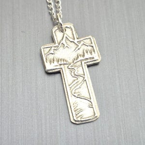 May include: Silver-toned pendant necklace featuring a cross design. The cross pendant has an embossed mountain and river scene. The necklace is suspended from a delicate silver chain, creating a simple and elegant accessory.