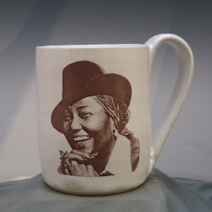 May include: White ceramic mug with a brown image of a woman wearing a hat and smiling. The woman is holding a cigarette in her hand.