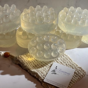 May include: Six clear round bath scrubbers with raised bumps, stacked on top of each other. A small white bag with a logo that reads "Accessories Beauty Suite" and the website address "www.accessoriesbeautysuite.com" and "Etsy: Accessories Beauty Suite" is in the foreground.