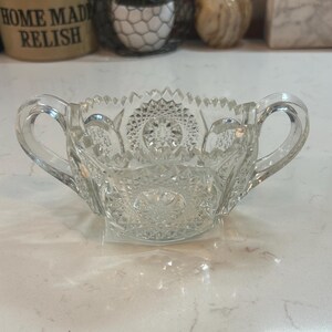 May include: A clear, cut-glass sugar bowl with two handles. The bowl has a detailed, starburst pattern on the bottom and around the rim. The handles are curved and attached to the sides of the bowl. A classic piece for serving sugar or other condiments.