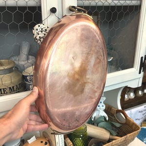 May include: A copper oval-shaped pan with a handle. The pan has a shiny, reflective surface and is held up in front of a white cabinet. The pan is likely used for cooking and is a vintage kitchen item.