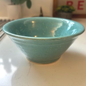 May include: A handmade, turquoise ceramic bowl with a speckled finish. The bowl has a rounded shape with a slightly flared rim and a smooth, glossy surface. The bowl is likely used for serving food or as a decorative piece.