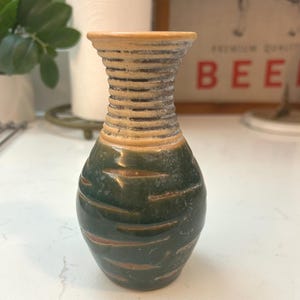 May include: A small, handcrafted ceramic vase with a textured, ribbed neck and a rounded body. The vase features a color palette of green, brown, and beige. The vase is suitable for displaying small flowers or as a decorative accent.