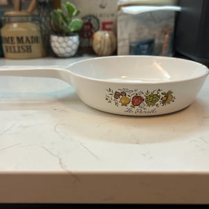 May include: White ceramic skillet with a single handle. The skillet features a colorful floral and vegetable design with the text "Le Persil" below the design. The skillet is on a white countertop.