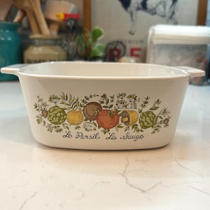 May include: White rectangular casserole dish with a colorful vegetable and herb design. The dish features illustrations of tomatoes, artichokes, and other produce, with the text "Le Persil La Sauge" below the design.