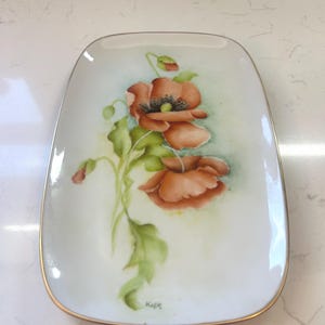 May include: Rectangular white ceramic dish with a gold rim, hand-painted with orange poppies and green leaves. The floral design is centered on the dish, with a signature in the lower right corner. The dish has rounded corners.