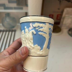 May include: A small, cream-colored ceramic cup with a light blue background and white relief figures. The cup has a black rim and base, and a ribbed bottom section. The design features a classical scene with trees and figures.
