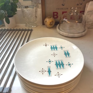 May include: A stack of round white ceramic plates featuring a repeating pattern of teal and brown geometric starburst and diamond shapes. The plates have a smooth, glossy finish and are arranged in a neat vertical pile on a flat surface.