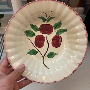 May include: Cream-colored ceramic plate with a scalloped edge and a hand-painted design of three red apples with green leaves. The rim has a distressed red border. A vintage-style decorative plate.