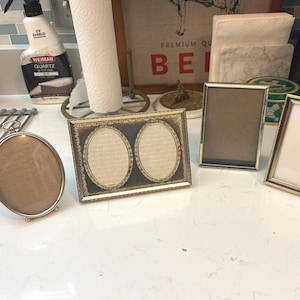 May include: A collection of vintage picture frames in various shapes and sizes. The frames include an oval frame, a double oval frame, and two rectangular frames. The frames have gold and silver finishes, with some featuring ornate detailing.
