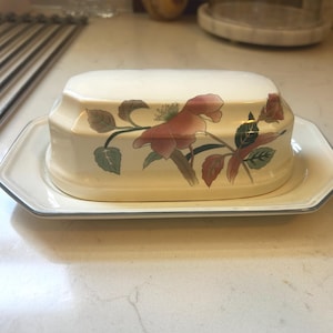 May include: A cream-colored ceramic butter dish with a rectangular base and a matching lid. The lid features a floral design with pink blossoms and green leaves. The edges of the base and lid are accented with a thin, dark gray or black trim line.