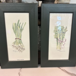 May include: Two framed botanical prints. One features a watercolor of green onions tied with a brown twine, the other shows a bunch of celery tied with a pink ribbon. Both are in dark green frames.