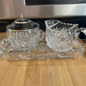 May include: A clear, cut-glass sugar bowl with a silver lid and a creamer sit on a matching tray. The sugar bowl and creamer have handles and are decorated with a starburst pattern. The tray is rectangular with a decorative edge.