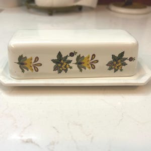 May include: A white ceramic butter dish with a lid, resting on a matching rectangular tray. The dish and lid are decorated with a floral pattern in yellow, green, and brown. The butter dish is a kitchenware item.