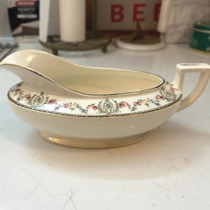 May include: Cream-colored gravy boat with a floral pattern around the rim and a gold trim. The gravy boat has a handle and a spout for easy pouring. A classic piece for serving sauces and gravies.
