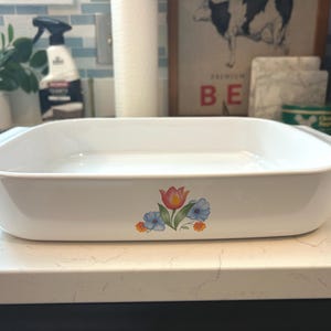 May include: A white rectangular baking dish with a floral design featuring a red and yellow tulip and blue flowers. The dish has handles on each side and is sitting on a white countertop.