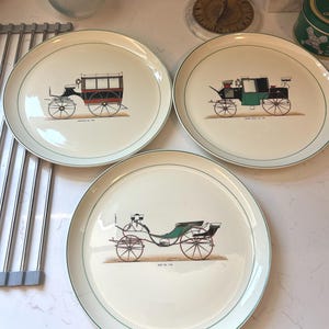 May include: Three cream-colored plates with a green border, each featuring a different vintage carriage illustration in shades of red, black, and green. The plates are arranged on a white countertop, showcasing a classic design.