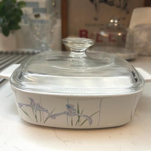 May include: A square white ceramic casserole dish with a clear glass lid and integrated side handles. The exterior features a delicate floral pattern of light purple iris flowers with green stems and leaves painted along the sides.