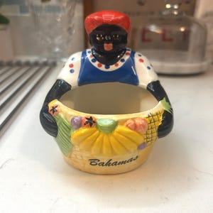 May include: A ceramic figurine bowl featuring a person wearing a red headscarf and a blue and white polka dot top. The person holds a yellow basket filled with colorful fruit, including yellow bananas. The word Bahamas is printed on the basket.