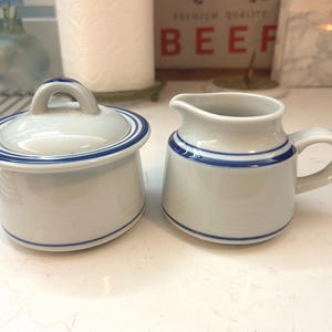 May include: A white ceramic sugar bowl with a lid and a small creamer. Both pieces have blue stripes around the rims. The sugar bowl has a handle on the lid. The creamer has a handle on the side.