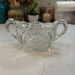 May include: Clear, cut-glass sugar bowl with two handles. The bowl features a detailed, geometric pattern and a scalloped rim. This vintage-style bowl is perfect for serving sugar or other condiments.