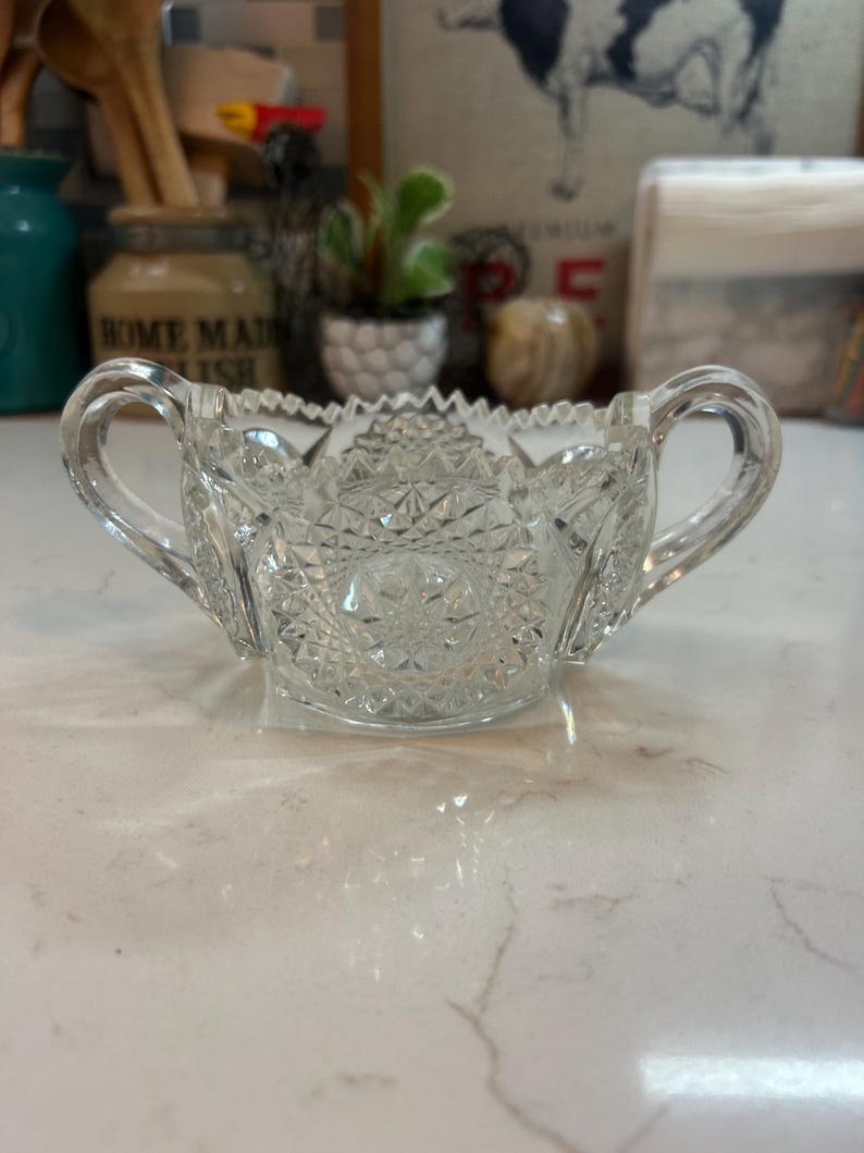 May include: Clear, cut-glass sugar bowl with two handles. The bowl features a detailed, geometric pattern and a scalloped rim. This vintage-style bowl is perfect for serving sugar or other condiments.
