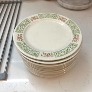 May include: A stack of round, off-white ceramic plates featuring a decorative border pattern. The rim is adorned with alternating green and brown scrollwork motifs arranged in rectangular segments around the edge of each plate.