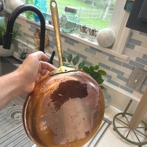 May include: A copper-bottomed saucepan with a brass handle. The pan has a rounded shape and a long handle with a hole for hanging. The copper exterior shows signs of wear, with areas of discoloration and patina.