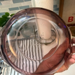 May include: A clear, round Pyrex glass bowl with a pinkish-purple hue. The bowl has a wide rim and a slightly curved base. The text "PYREX" is visible on the bottom, along with other text. The bowl is held by a hand.