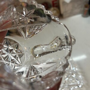 May include: Clear crystal glass with starburst and geometric patterns. The glass has a cut design with a textured surface. The image shows a close-up of the glass, highlighting the intricate details and clarity of the material.