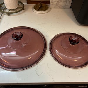 May include: Two matching, round, purple glass lids with dark purple knobs. The lids have a slightly raised, textured design around the knob. One lid is larger than the other. Kitchenware.