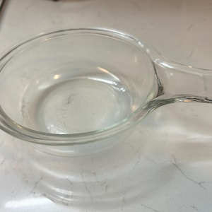 May include: A clear glass bowl with a handle. The bowl is round with a slightly flared rim and a pouring spout. The bowl is sitting on a white surface.