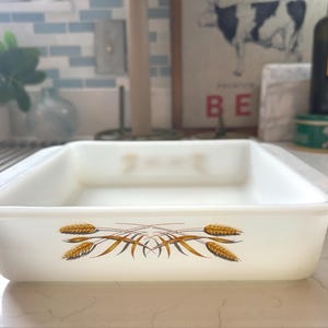 May include: A rectangular, white baking dish with a wheat stalk design in shades of brown and gold. The dish is made of a thick, opaque material and has a slightly rounded edge. The dish is sitting on a white countertop.