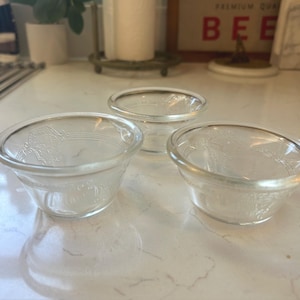May include: Three clear glass bowls with a decorative floral pattern. The bowls are small, with a rounded shape and a slightly flared rim. They are arranged on a light-colored surface.