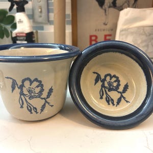 May include: Two ceramic bowls with a cream-colored base and a blue floral design. The rims are painted blue. One bowl is upright, and the other is on its side, revealing the interior floral pattern.