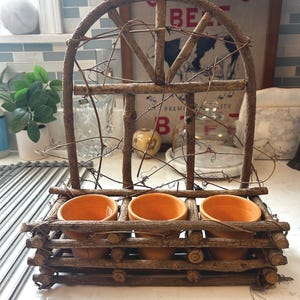 May include: A decorative planter stand constructed from natural brown twigs and branches, featuring an arched window-like frame and a rectangular base holding three small, empty, round terracotta-colored clay pots in a row.