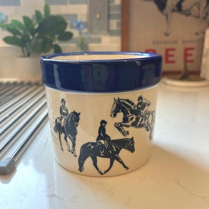 May include: A cream-colored ceramic container with a navy blue rim. The container features black illustrations of equestrian figures on horseback. The container is sitting on a white countertop.
