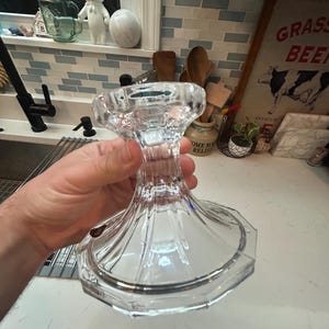 May include: A clear, faceted glass candle holder with a wide base and a flared top. The candle holder is held in a hand, showcasing its elegant design. The kitchen background includes a sink, window, and decorative items.