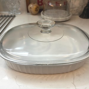 May include: An oval white ceramic baking dish with vertical fluted sides and a clear glass lid featuring a central knob handle. The dish has a smooth, glossy finish and a transparent cover that allows for a clear view of the interior base.