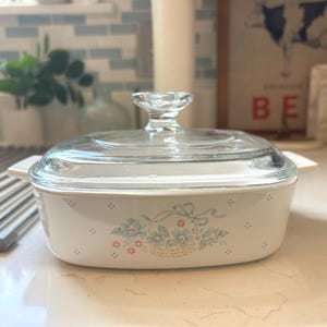 May include: A white casserole dish with a clear glass lid. The dish features a floral design with blue and pink flowers and a basket. The handles are white. The dish is on a countertop.