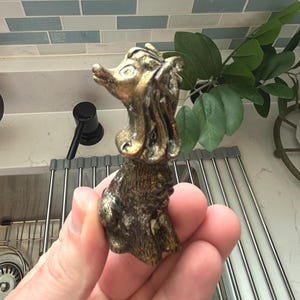 May include: A small, antique-style dog figurine with a gold and bronze finish. The dog is posed upright, with detailed fur and facial features. The figurine is held in a hand.